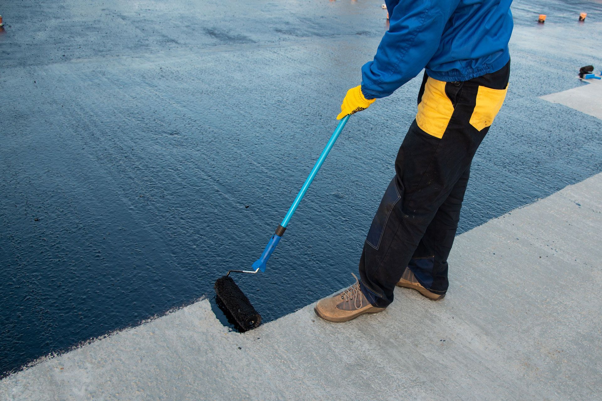 Person in blue jacket and yellow gloves rolling black coating onto a concrete surface with a paint roller