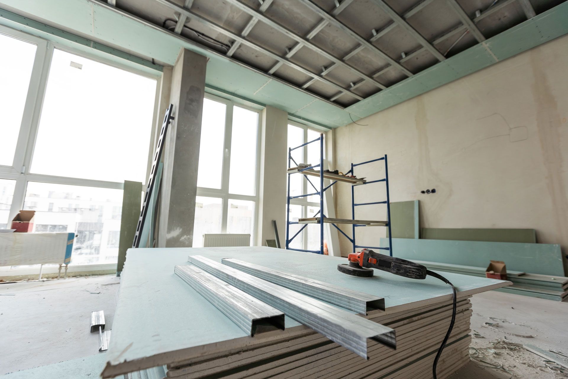 Interior of a room under construction with stacked drywall sheets, scaffolding, and large windows.