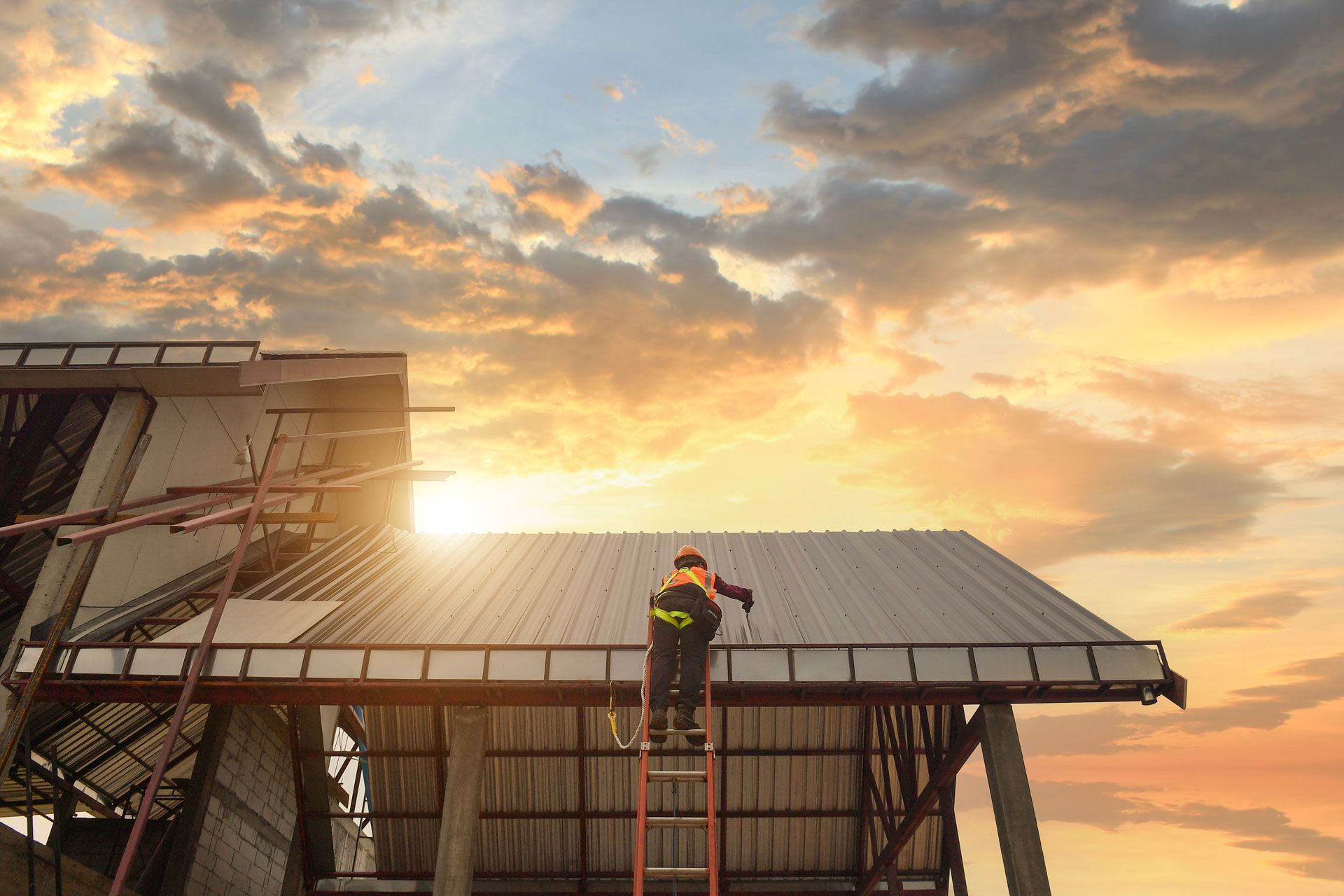 Worker on a ladder installing a metal roof at sunset under a cloudy sky