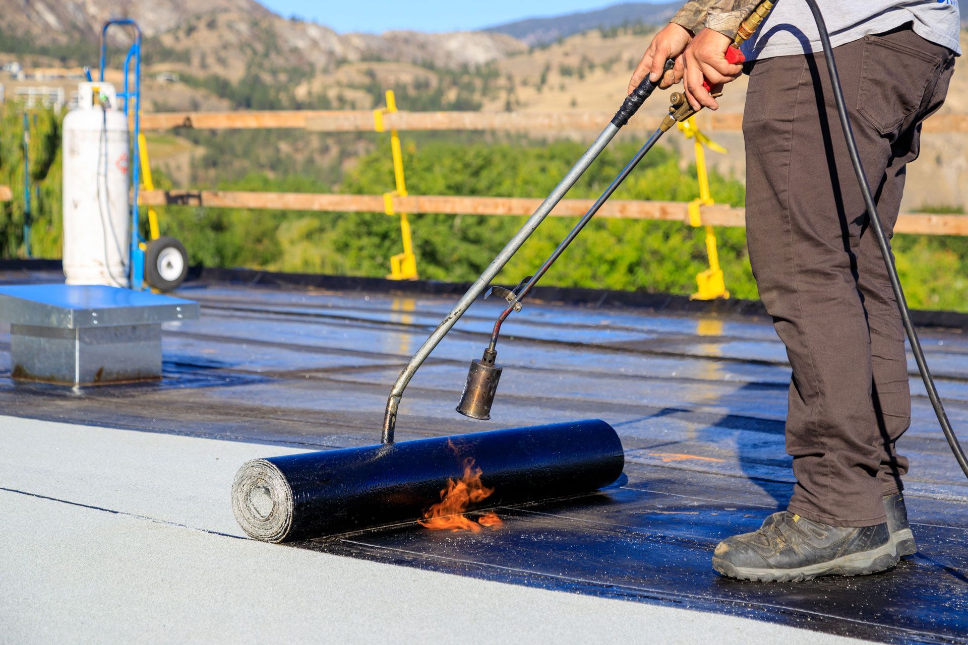 Person torching a flat roof membrane beside a bucket and roof tools outdoors