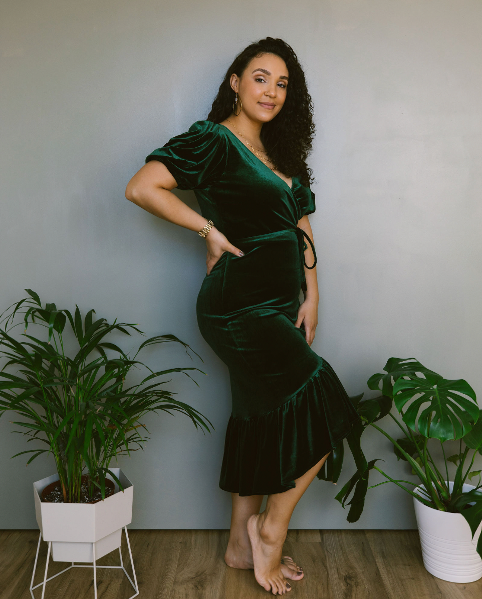 A woman in a green velvet dress is standing in front of potted plants.