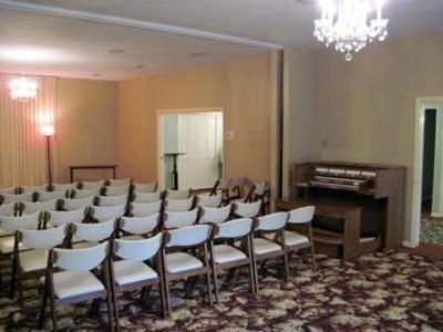 Small banquet room with rows of white chairs facing a lectern and upright piano on patterned carpet.