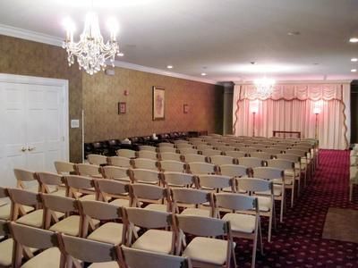 Empty banquet hall with rows of beige chairs facing a small stage with pink curtains and chandeliers