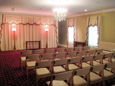 Empty banquet hall with rows of chairs facing a draped stage and chandelier lighting