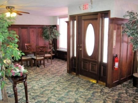 Hotel lobby hallway with wood-paneled walls, patterned carpet, chairs, plants, and a glass-paneled door