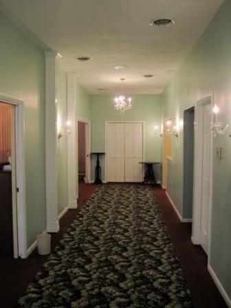 Dim hallway with patterned carpet, white walls, chandeliers, and closed double doors at the end.