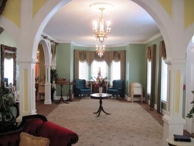 Elegant sitting room with arched columns, chandeliers, carpet, and teal chairs by the windows.