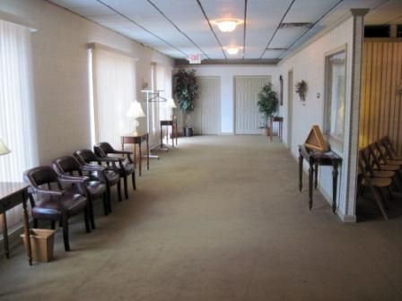 Empty waiting room with chairs along the walls, carpeted floor, and potted plants near the back.