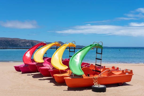 Colorful pedal boats lined up on a sandy beach under a blue sky and calm sea.