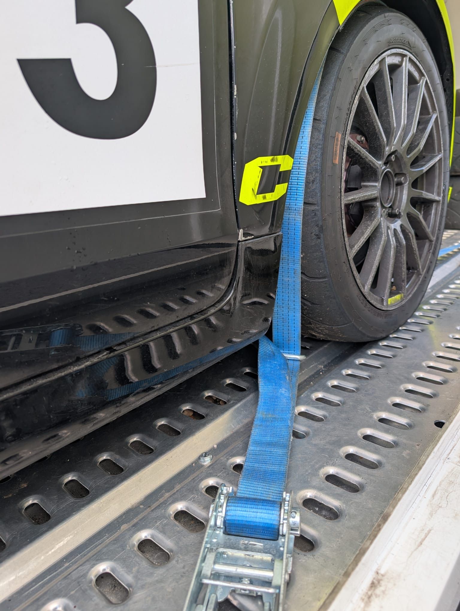 Blue strap securing a black race car's tire to a metal trailer bed. The car has a number 3 on its side.