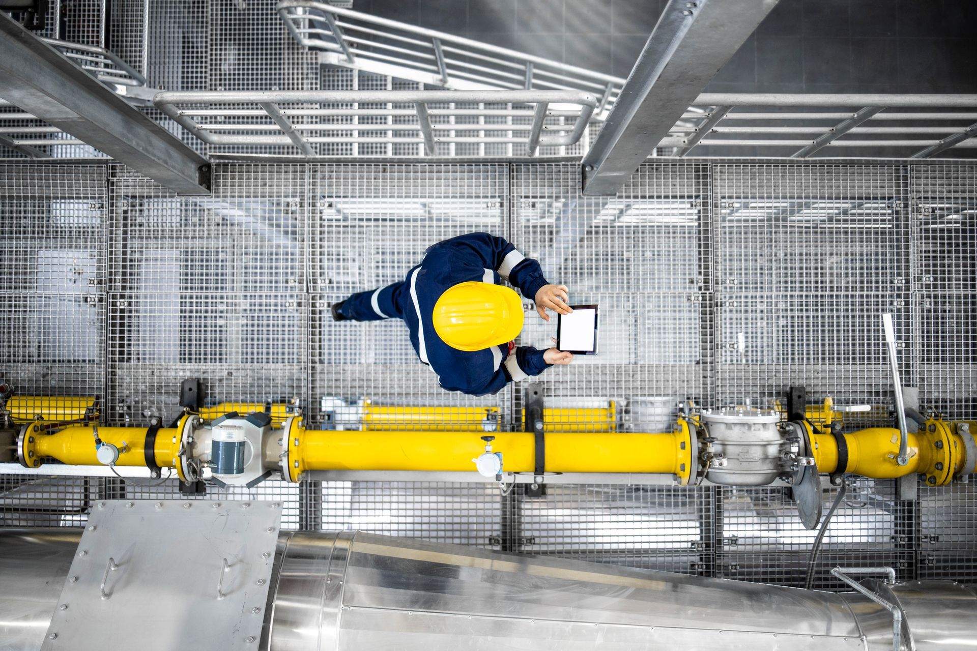 Engineer in yellow hard hat and blue uniform inspects industrial equipment with tablet.