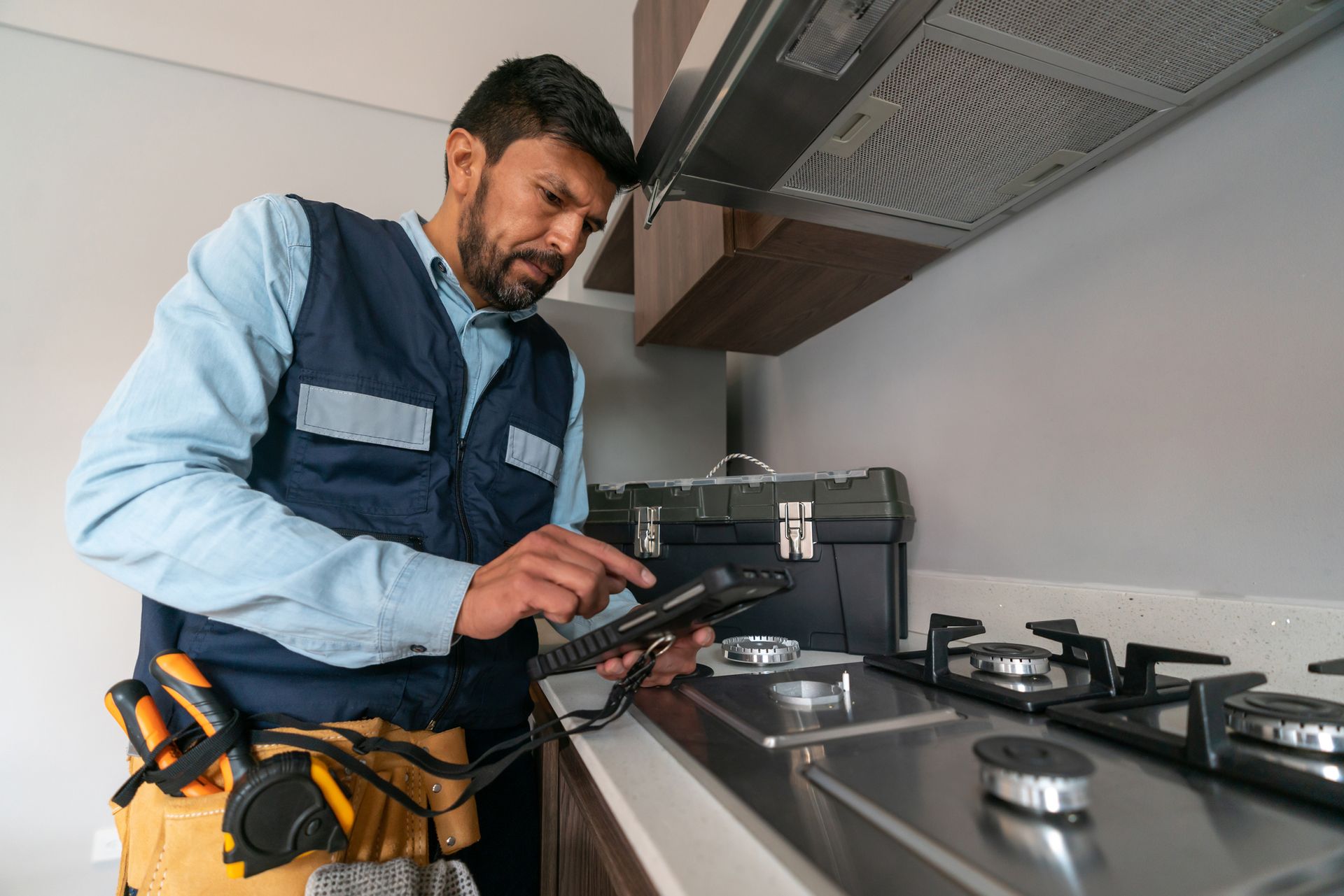 A repairman in blue vest examines a tablet near a stove in a kitchen..