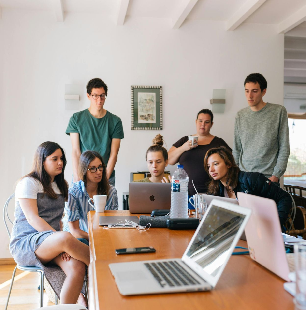 Group of people gathered around laptops in a bright meeting room, working together at a table.
