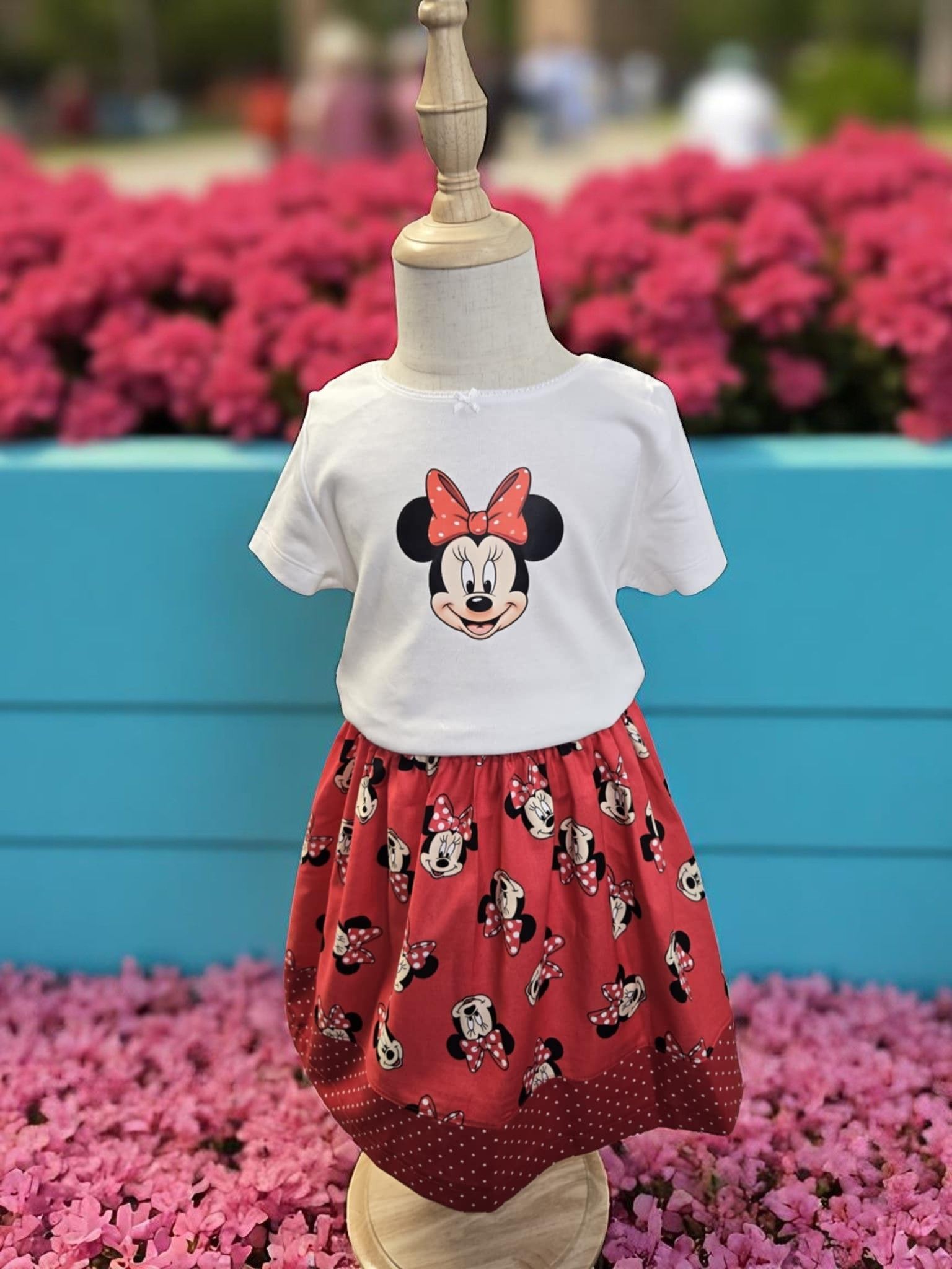 A white Minnie Mouse t-shirt and a red patterned Minnie Mouse skirt on a mannequin in front of pink flowers.
