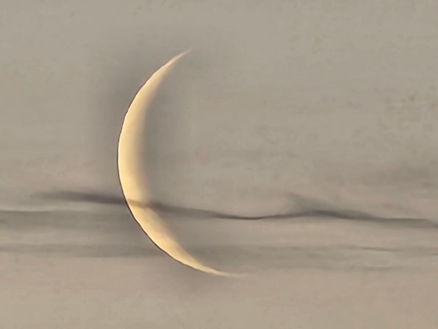 A thin crescent moon appearing against a backdrop of soft, hazy gray clouds.