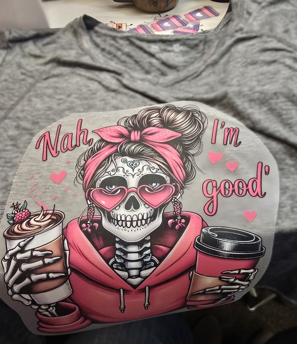 A gray t-shirt with a graphic of a skeleton wearing a pink hoodie, headband, and heart glasses, holding two coffee cups.