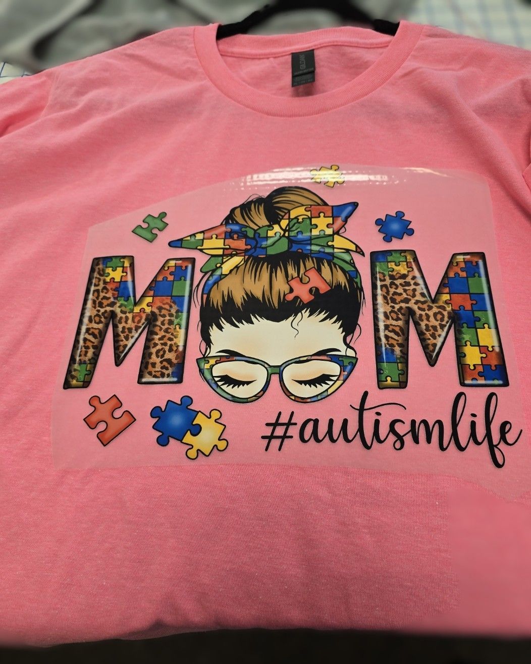A pink T-shirt with a graphic of a woman with a messy bun, glasses, and puzzle piece patterns, reading 
