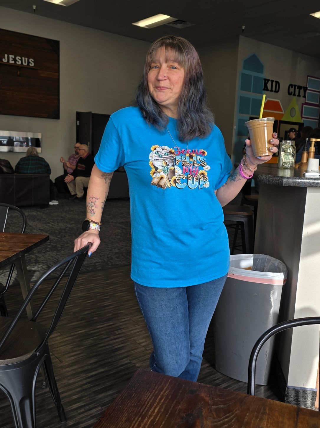 A smiling person wears a blue t-shirt and jeans, standing in a room while holding a plastic cup with a straw.