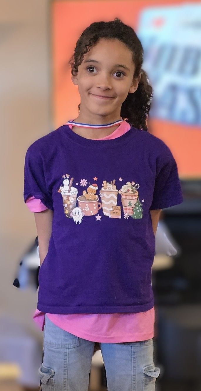 A person wears a purple t-shirt with a graphic of holiday drinks over a pink shirt, standing in an indoor setting.