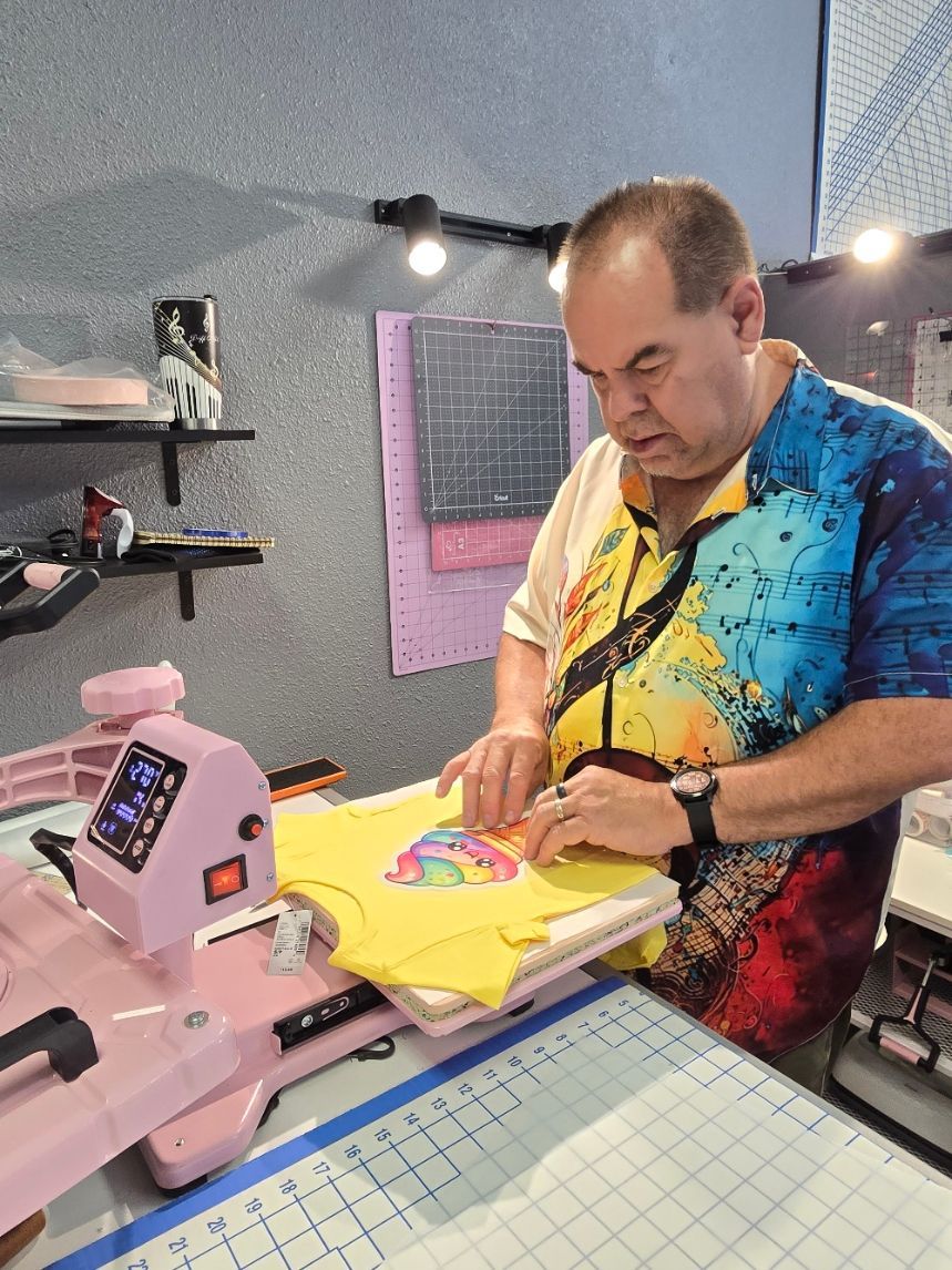A person wearing a colorful shirt uses a pink heat press to apply a design to a yellow t-shirt in a craft workspace.