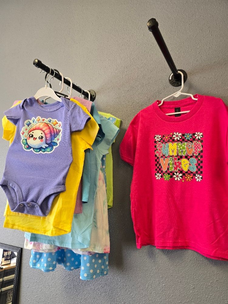 Children's clothing hangs on two wall-mounted racks: a purple bodysuit and several shirts in yellow, blue, and hot pink.