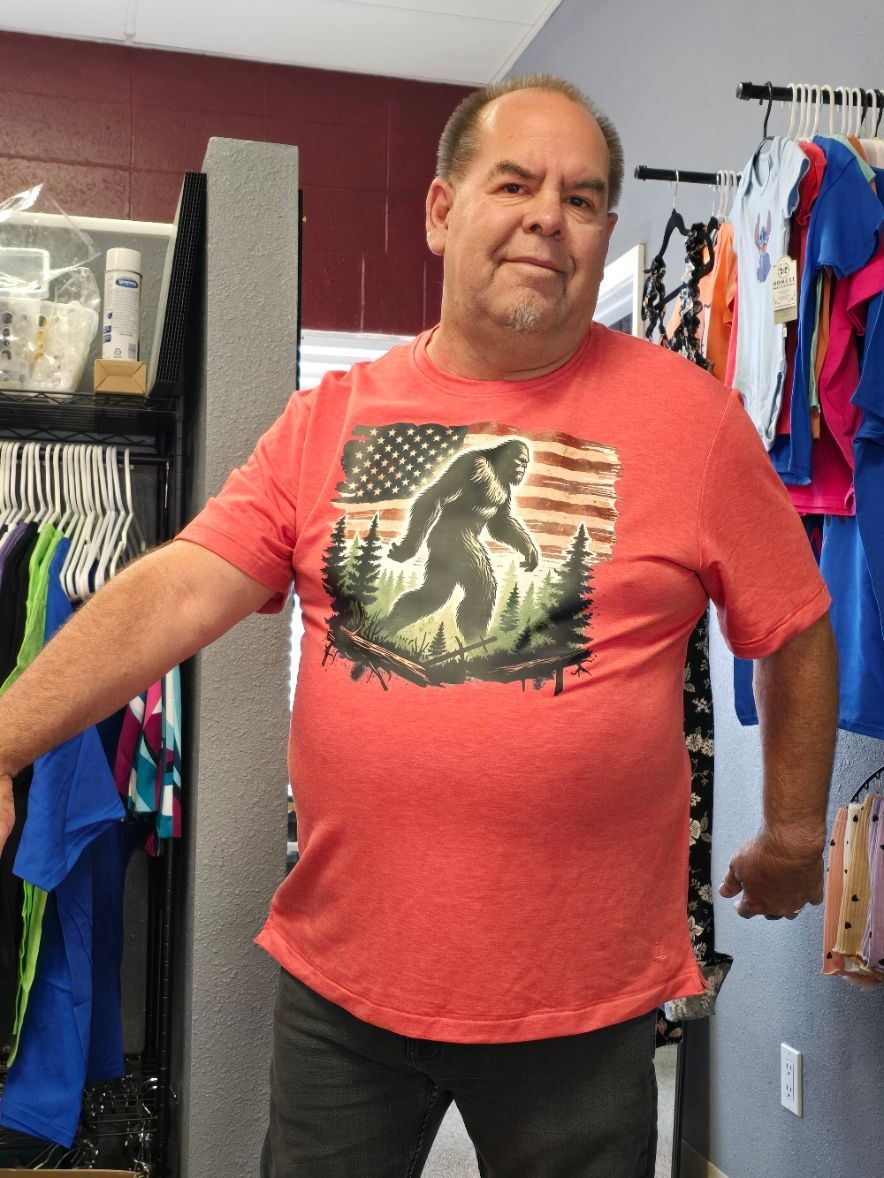 A person stands in a clothing shop wearing a coral-colored t-shirt featuring a silhouette of Bigfoot against a US flag.