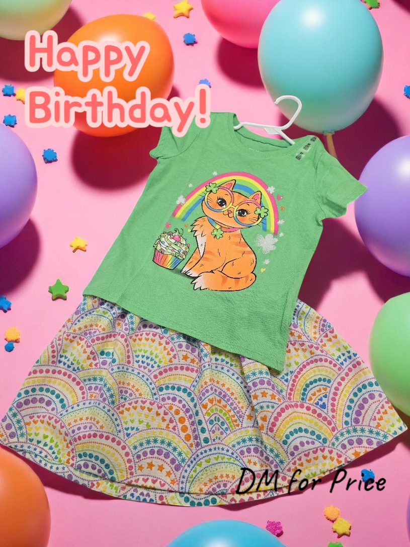 A green shirt with an orange cat and rainbow print over a rainbow-patterned skirt, surrounded by balloons and confetti.