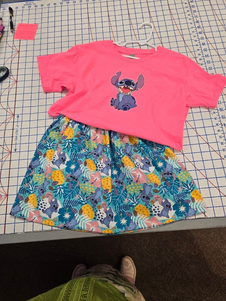 A bright pink t-shirt featuring a small Stitch graphic, paired with a patterned skirt on a grid-lined surface.