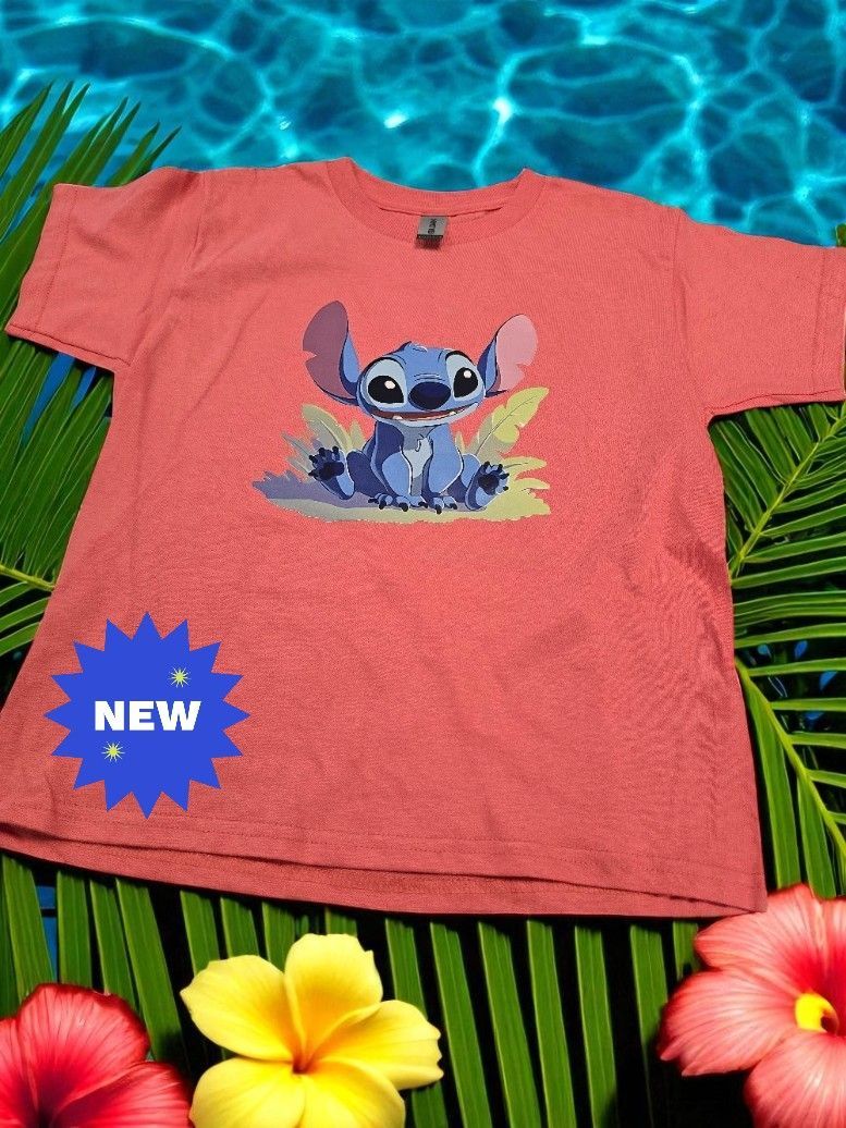 A coral-colored t-shirt featuring a Stitch graphic, displayed on a tropical background with palm leaves and flowers.