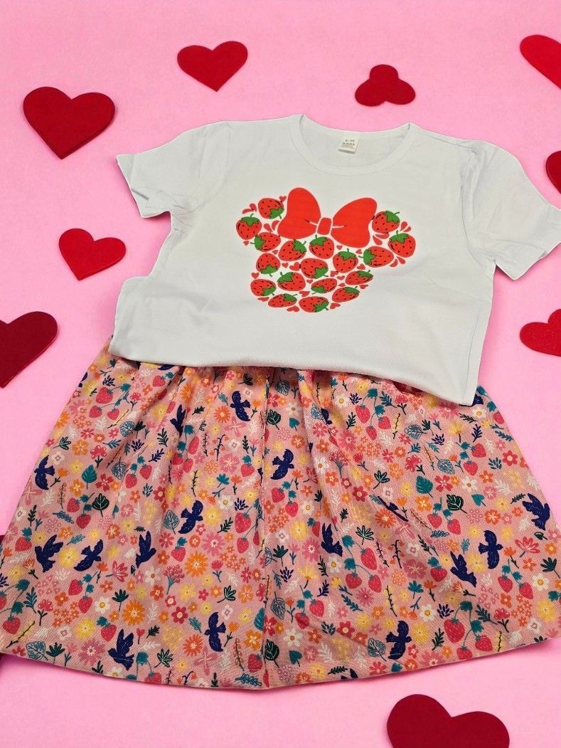 A white short-sleeved top with a strawberry-themed Minnie Mouse icon, paired with a floral skirt on a pink heart background.