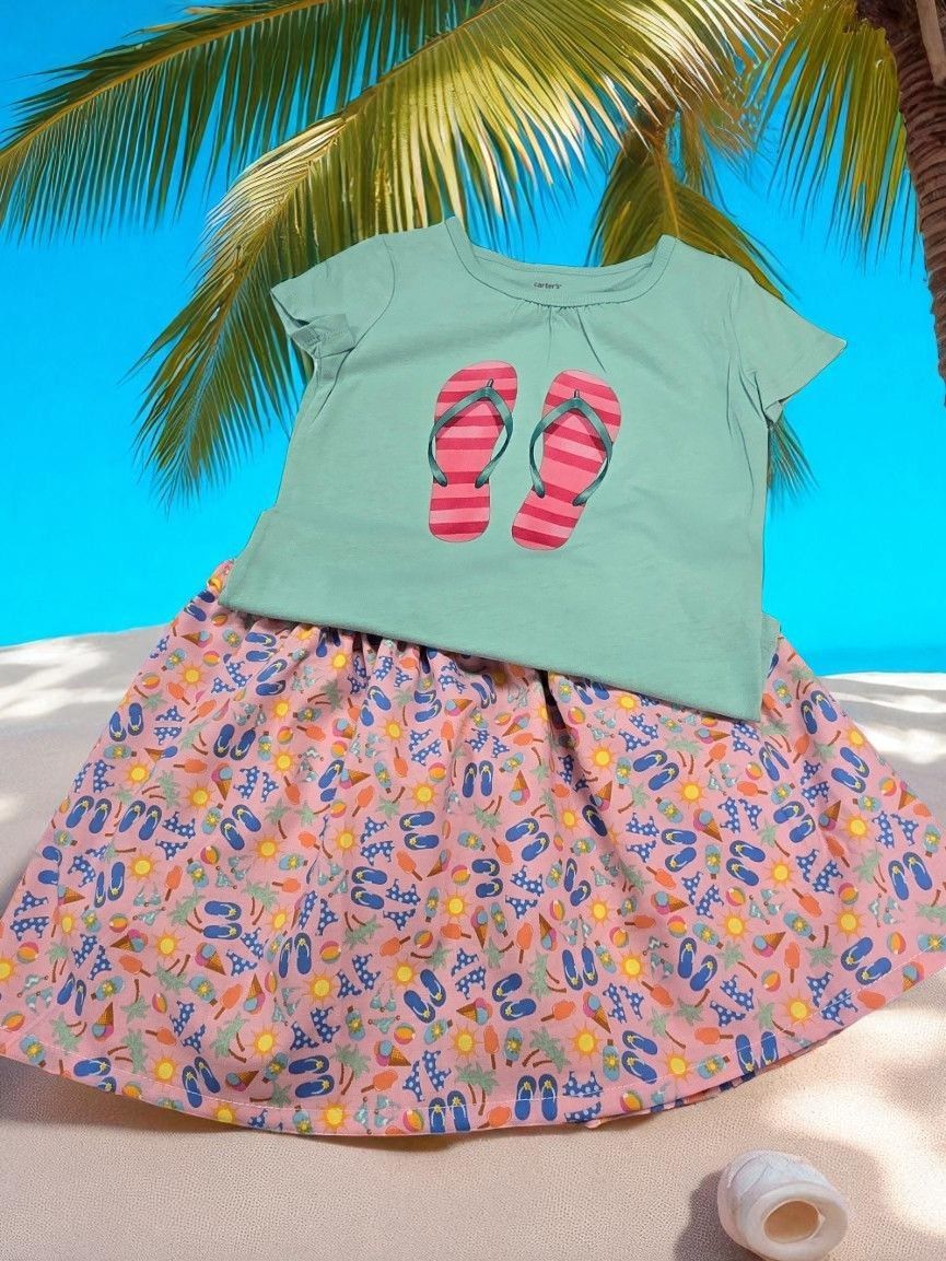 A mint-green t-shirt with a pink flip-flop print and a matching patterned skirt, displayed on a sandy beach.