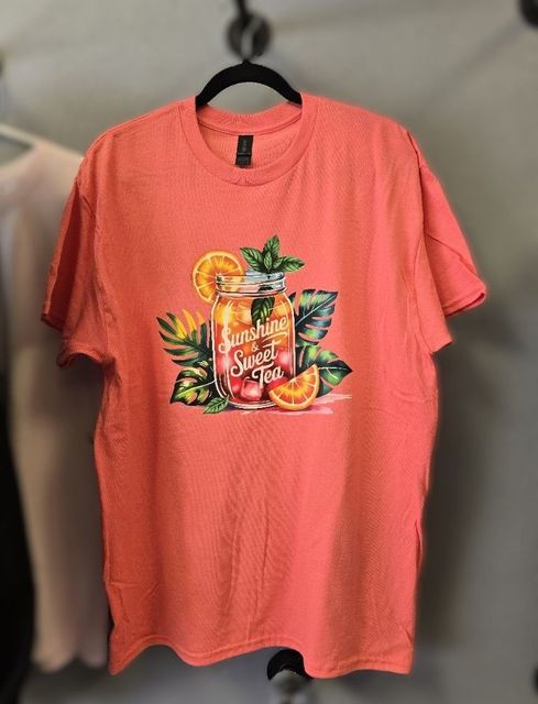 A coral-colored t-shirt featuring a graphic of a mason jar filled with sweet tea, orange slices, and tropical leaves.