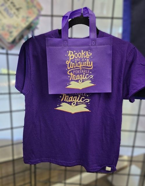 A purple t-shirt and matching tote bag hanging on a grid wall. Both feature the gold text "Books: A Uniquely Portable Magic."