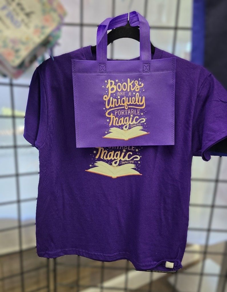 A purple t-shirt and matching tote bag hanging on a grid display, both featuring the golden text 