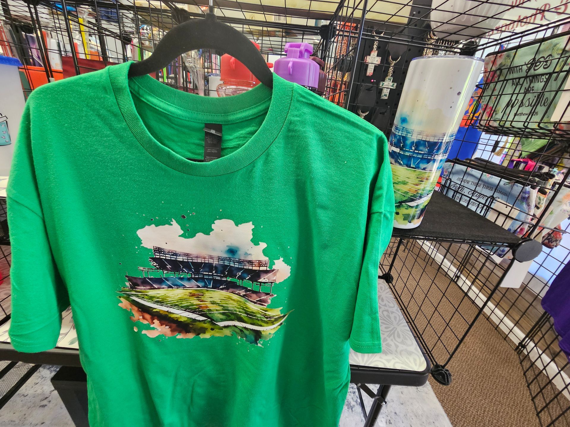 A bright green t-shirt featuring a watercolor-style illustration of a stadium, displayed in a retail setting.