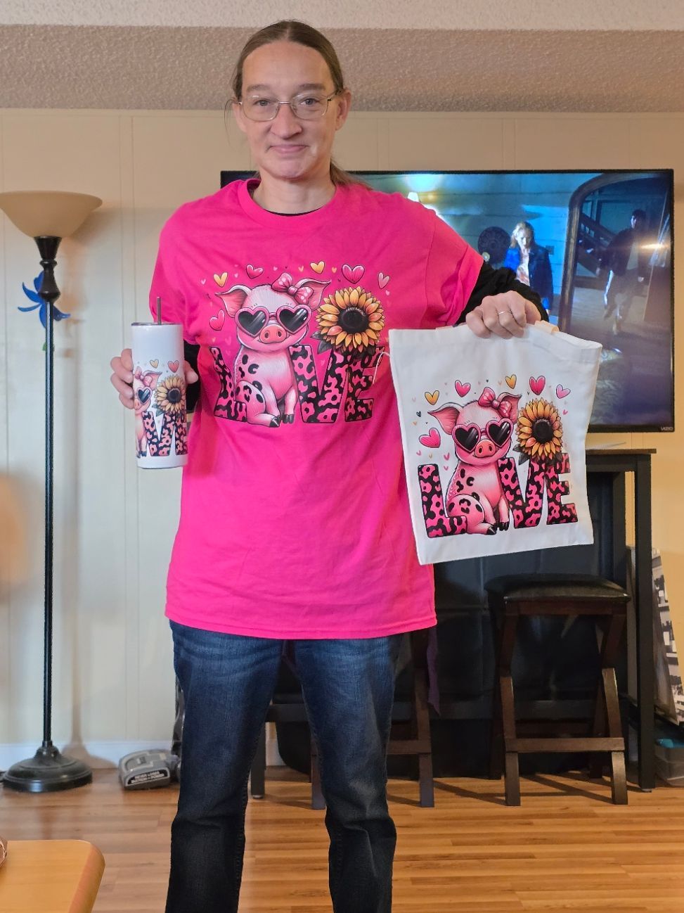 A person stands in a living room holding a matching tumbler and fabric square featuring a pig, sunflower, and 