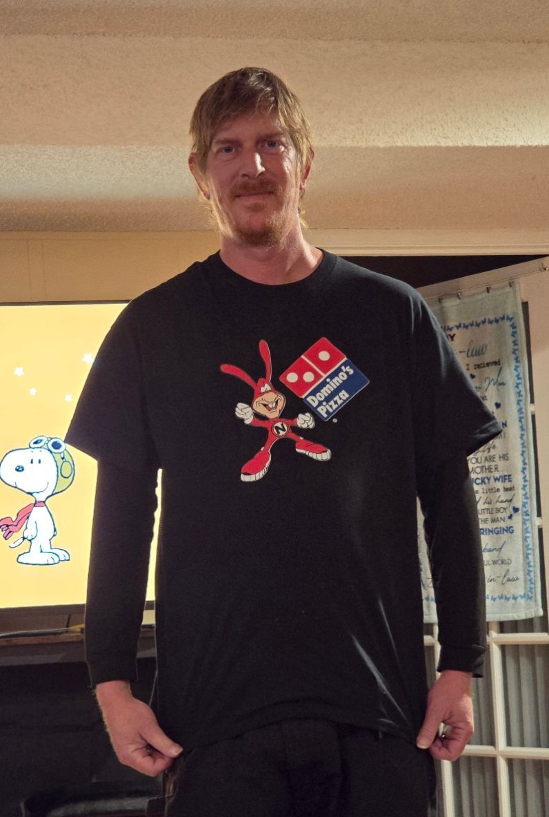A person stands indoors wearing a black t-shirt with a cartoon rabbit and Domino's Pizza logo, with a Snoopy display behind.