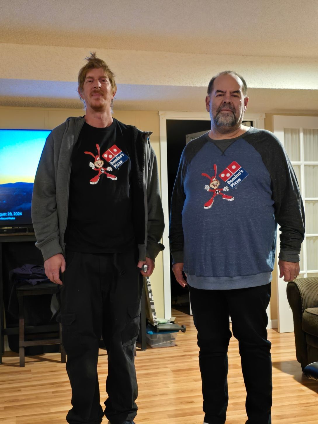 Two people stand indoors wearing matching Domino's Pizza Noid shirts, one with a grey hoodie and one with a blue shirt.