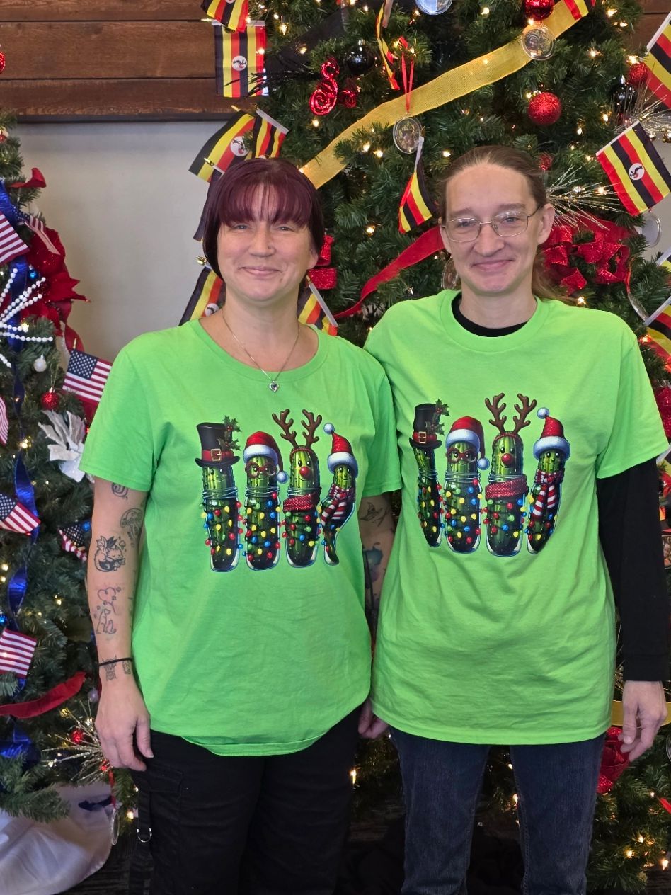 Two people in matching neon green Christmas pickle shirts stand in front of decorated holiday trees.