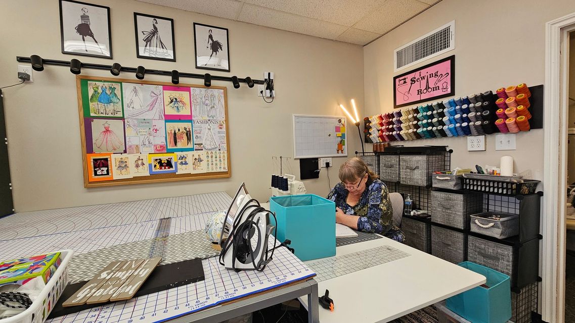 A person works at a desk in a craft room featuring a bulletin board, wall-mounted thread racks, and organized storage.