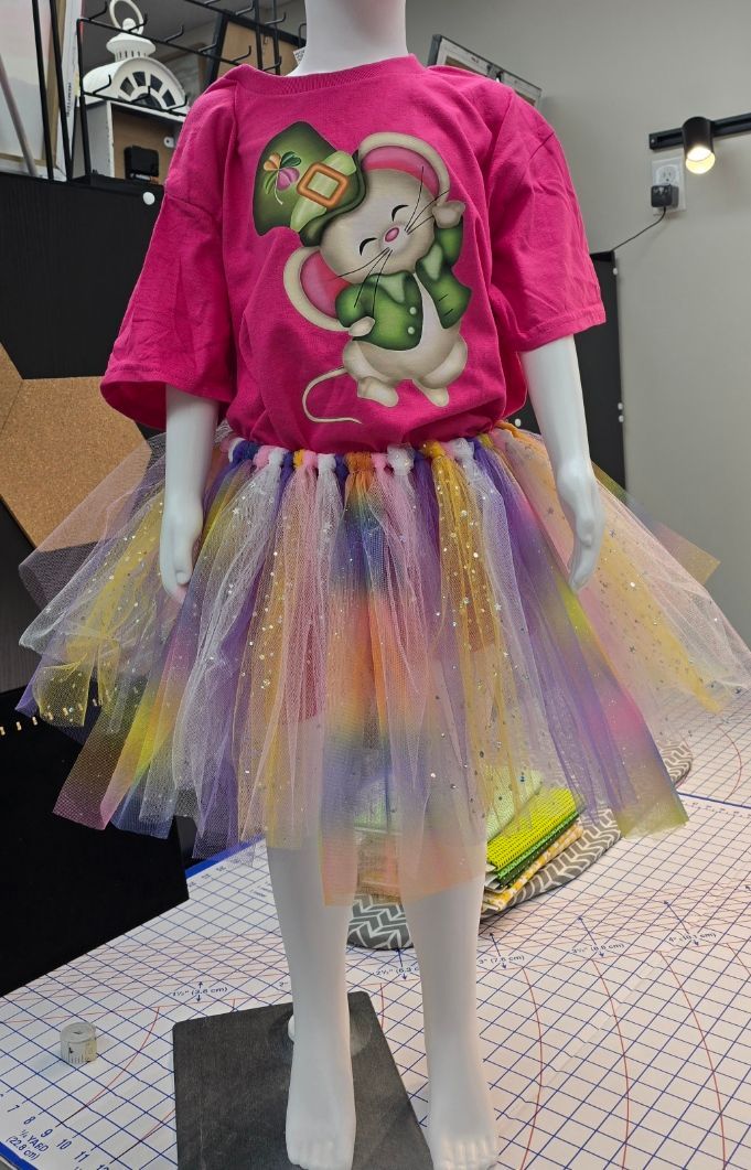 A mannequin wears a bright pink t-shirt with a mouse graphic and a colorful, sparkly tulle tutu.