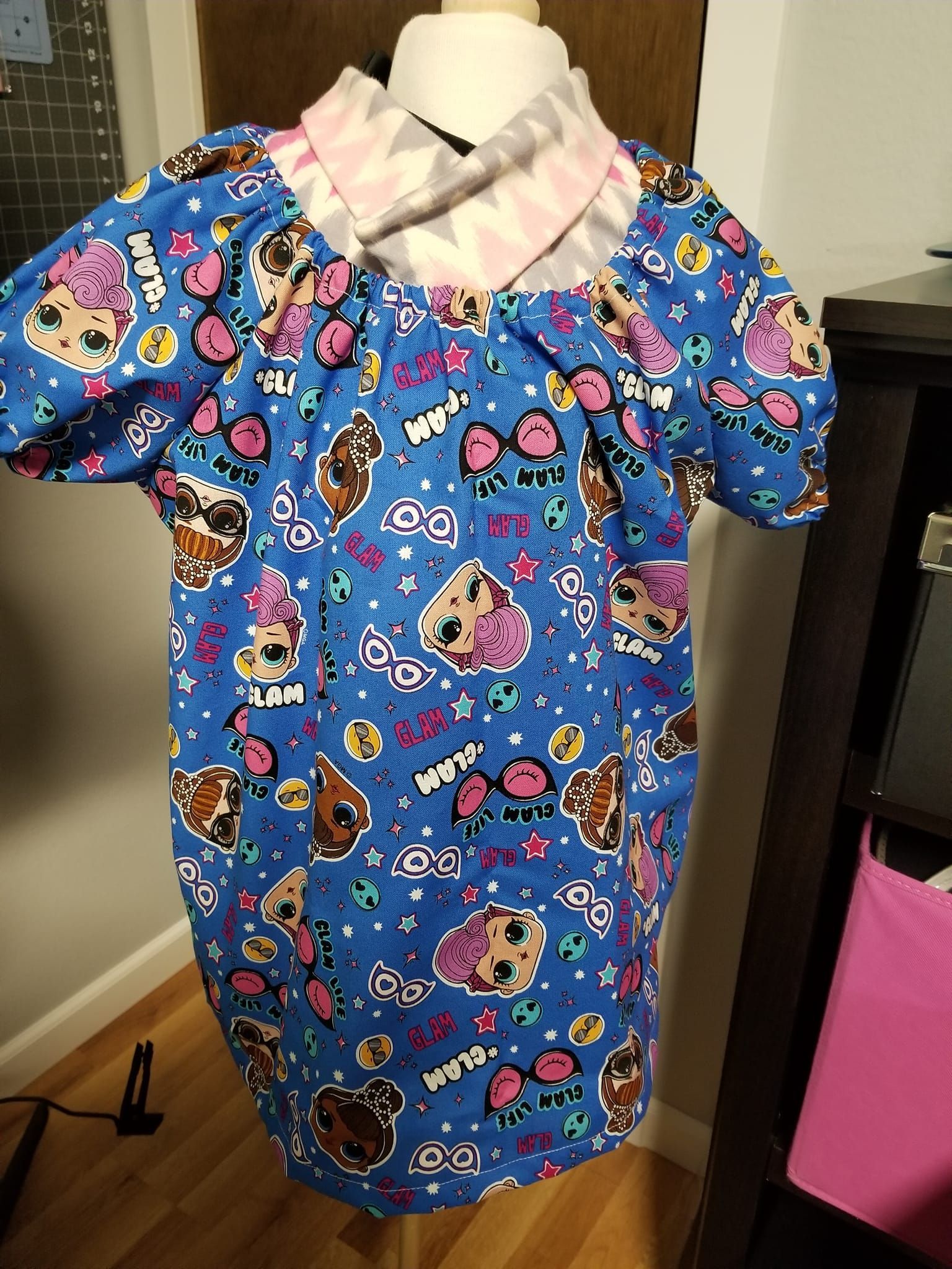 A child’s blue shirt featuring colorful doll and icon patterns, with a pink and white chevron-patterned folded collar.