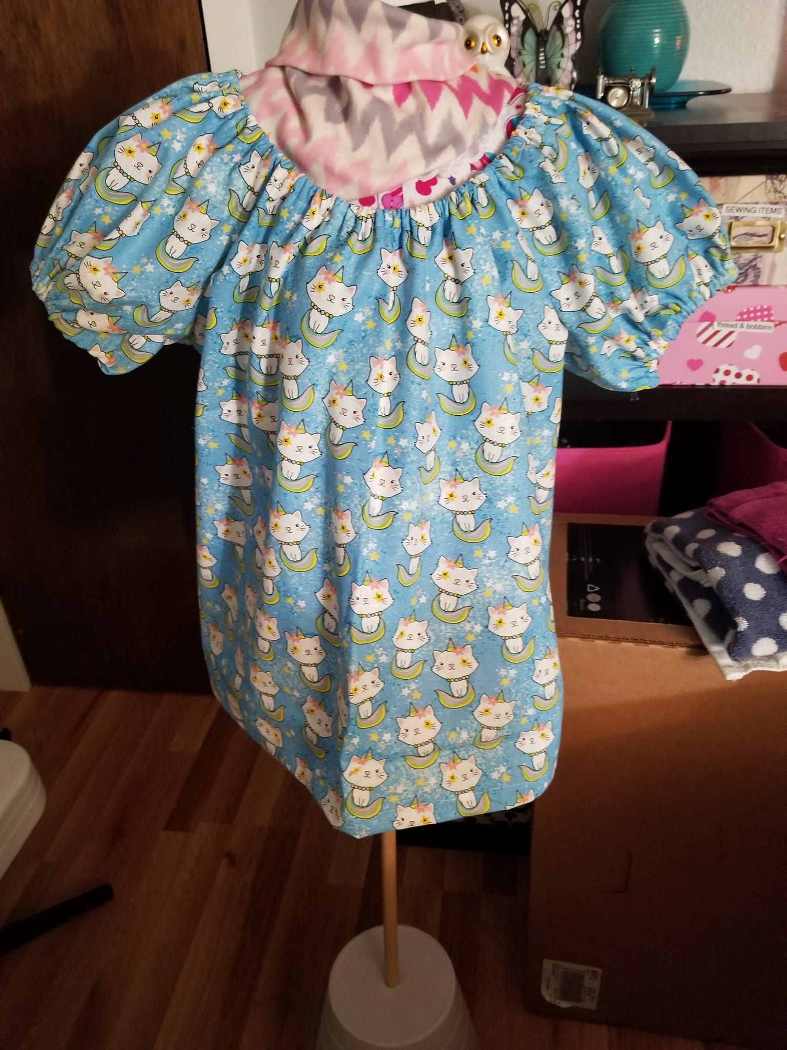 A light blue shirt with a cartoon cat print, puffed sleeves, and a gathered neckline displayed on a dress form.