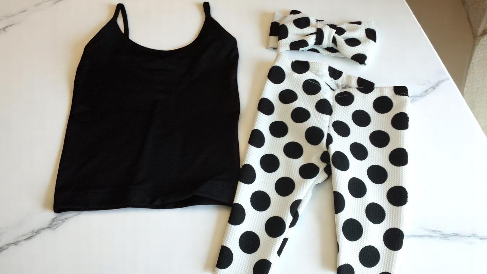 A black camisole top, white leggings with black polka dots, and a matching polka-dot headband on a white marble surface.