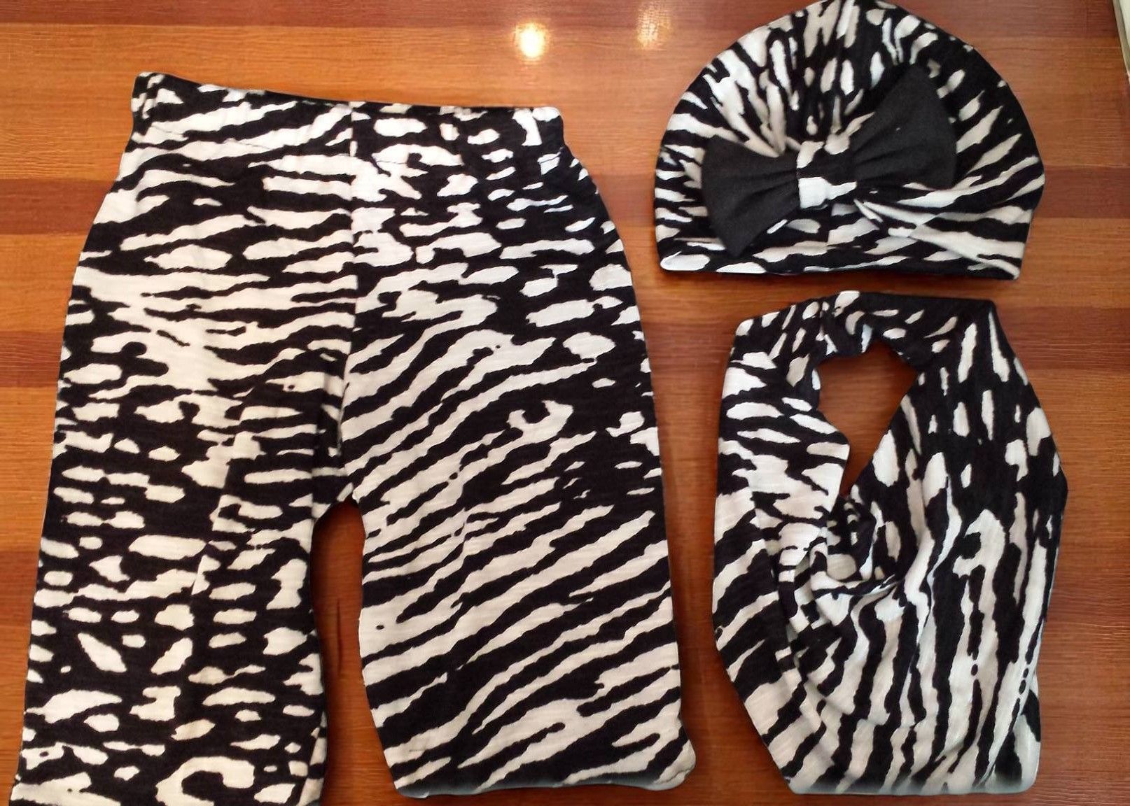 A set of children's clothing in a black-and-white animal print, featuring leggings, a bow-accented hat, and a scarf.