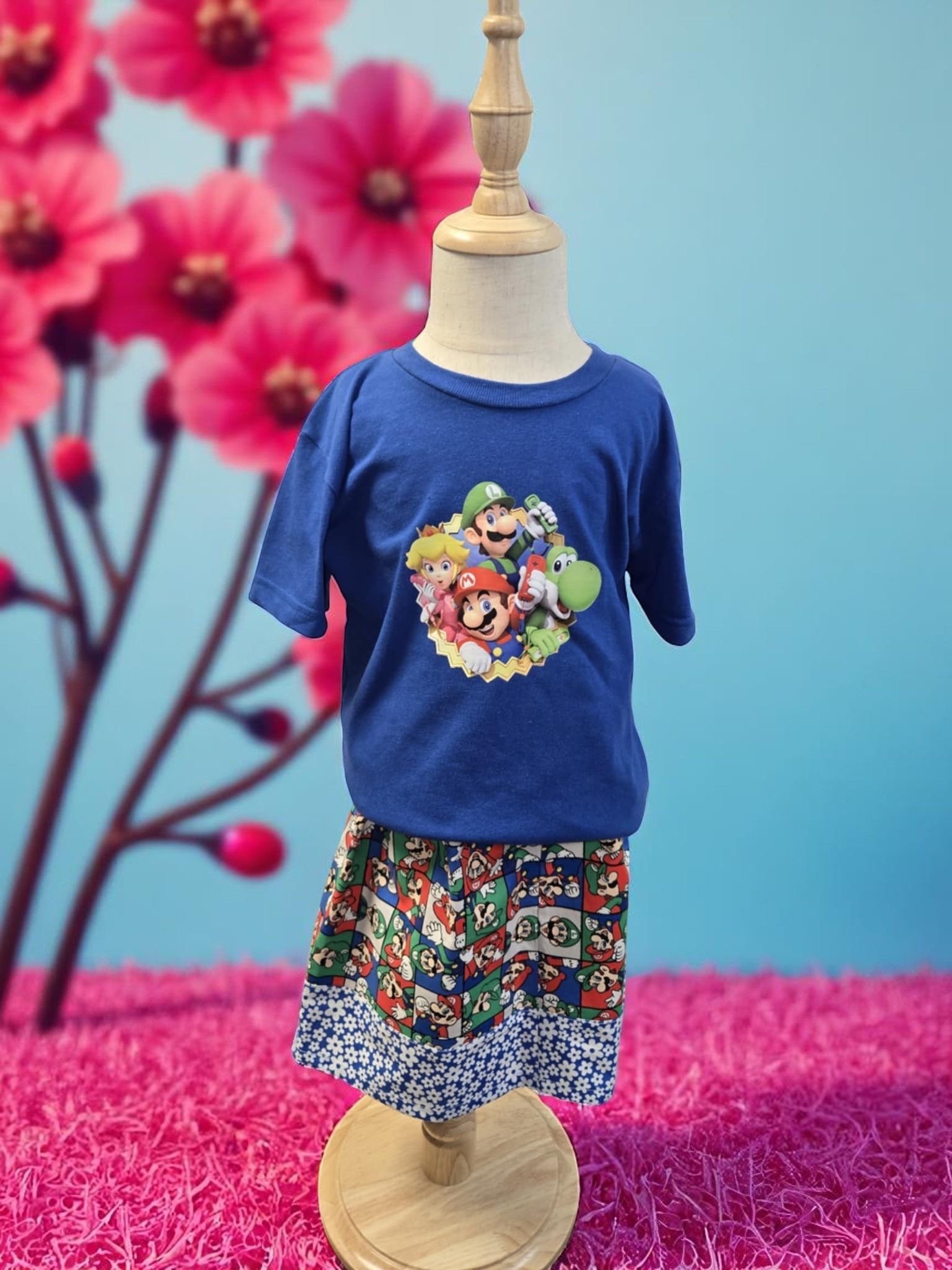 A blue Super Mario t-shirt and patterned shorts displayed on a child-sized mannequin in front of large pink flowers.