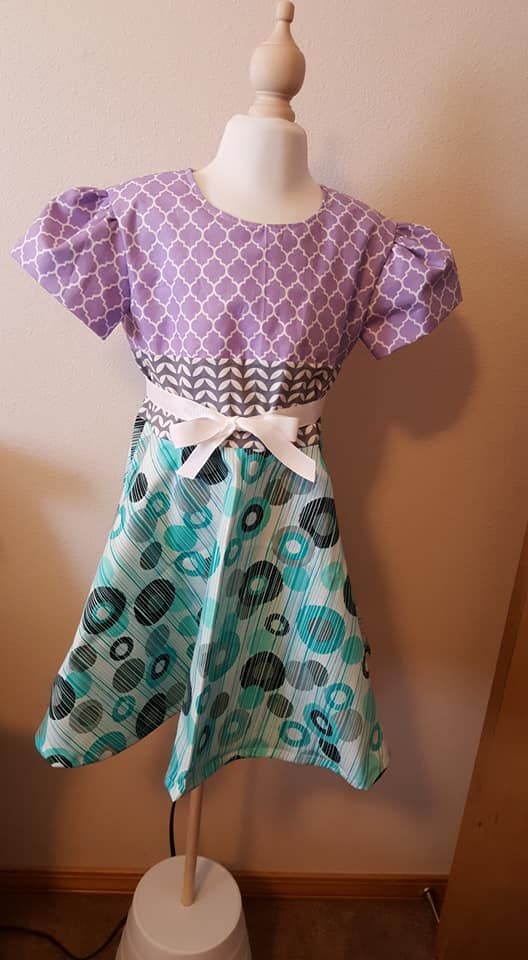A girl's dress on a mannequin with a purple scale-pattern bodice, chevron waistband, white bow, and blue circle-print skirt.