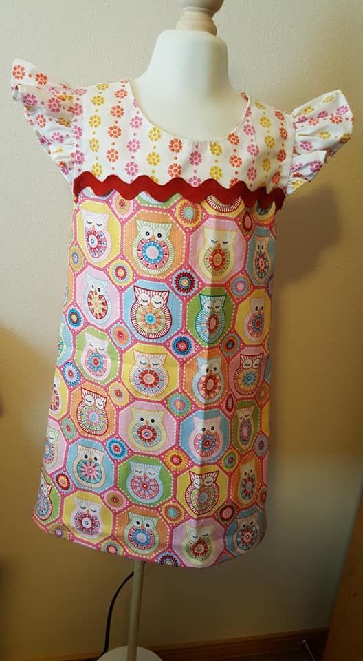 A girl's dress with a yellow floral bodice, flutter sleeves, red rickrack trim, and a colorful owl-patterned skirt.