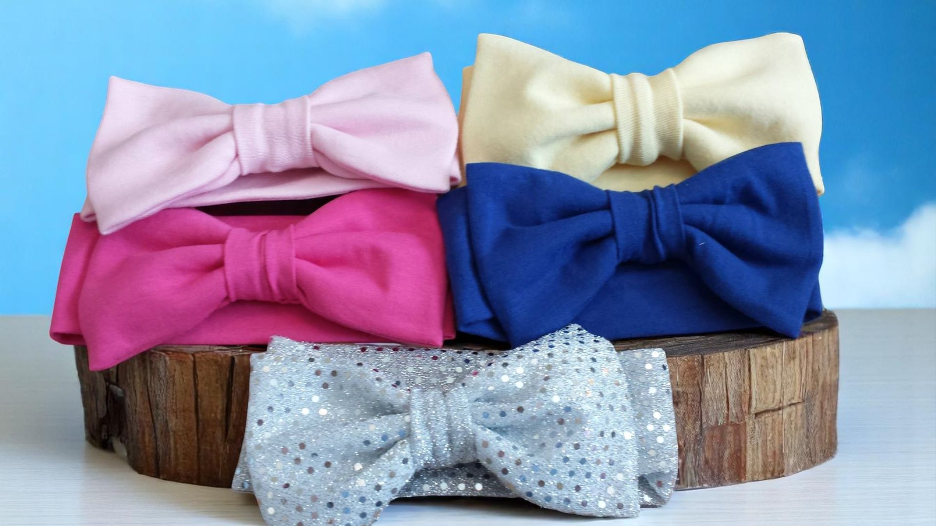 Five fabric hair bows in pink, hot pink, cream, blue, and a silver sequined style, arranged on a wooden log base.