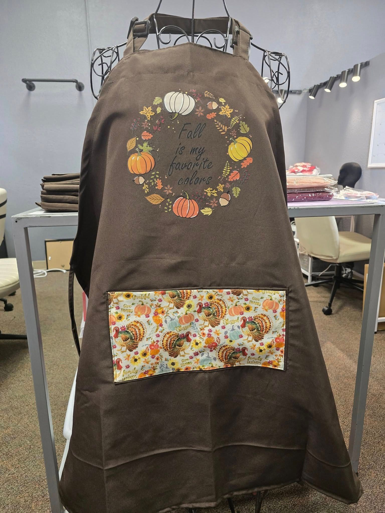 A brown apron on a display stand, featuring an embroidered autumn wreath design and a patterned pocket.
